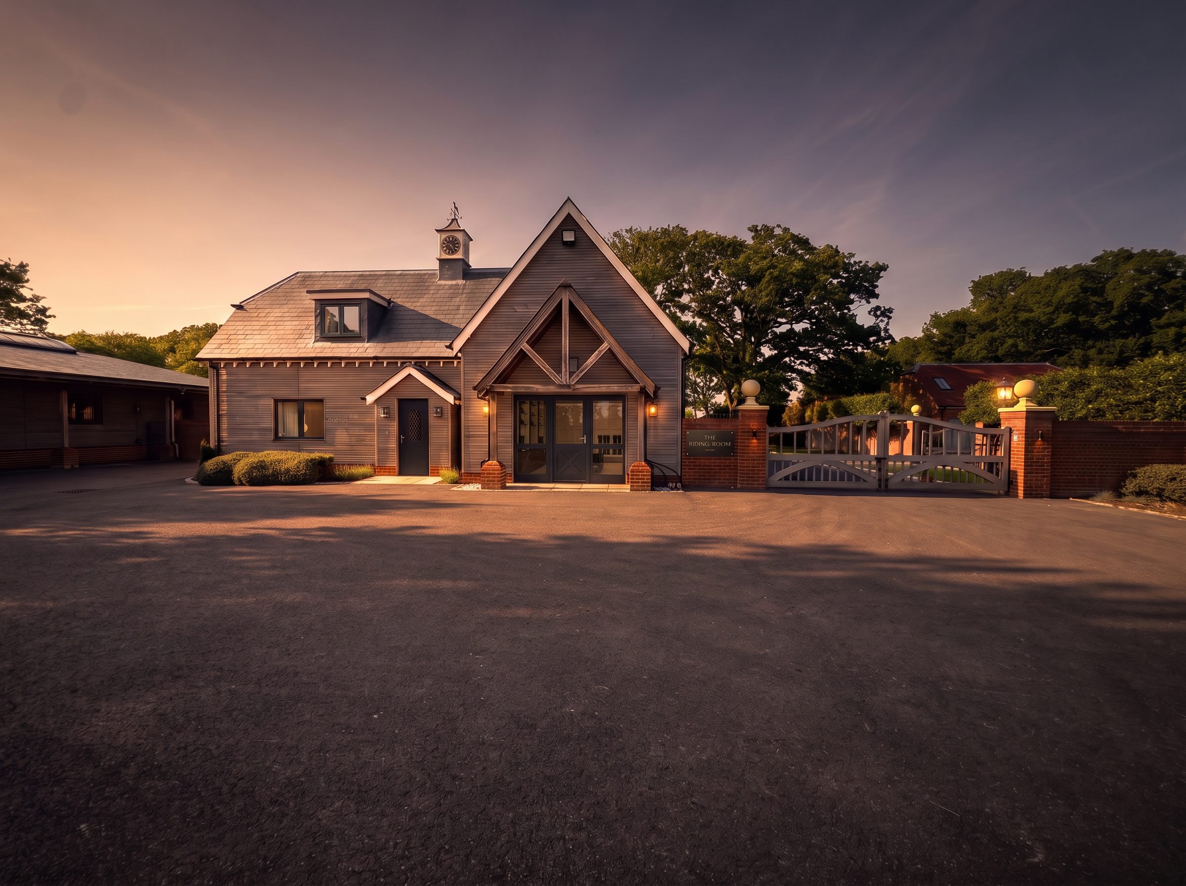 The Riding Room at Forest Oaks: a timber-clad building with a clock tower, warmly lit at dusk